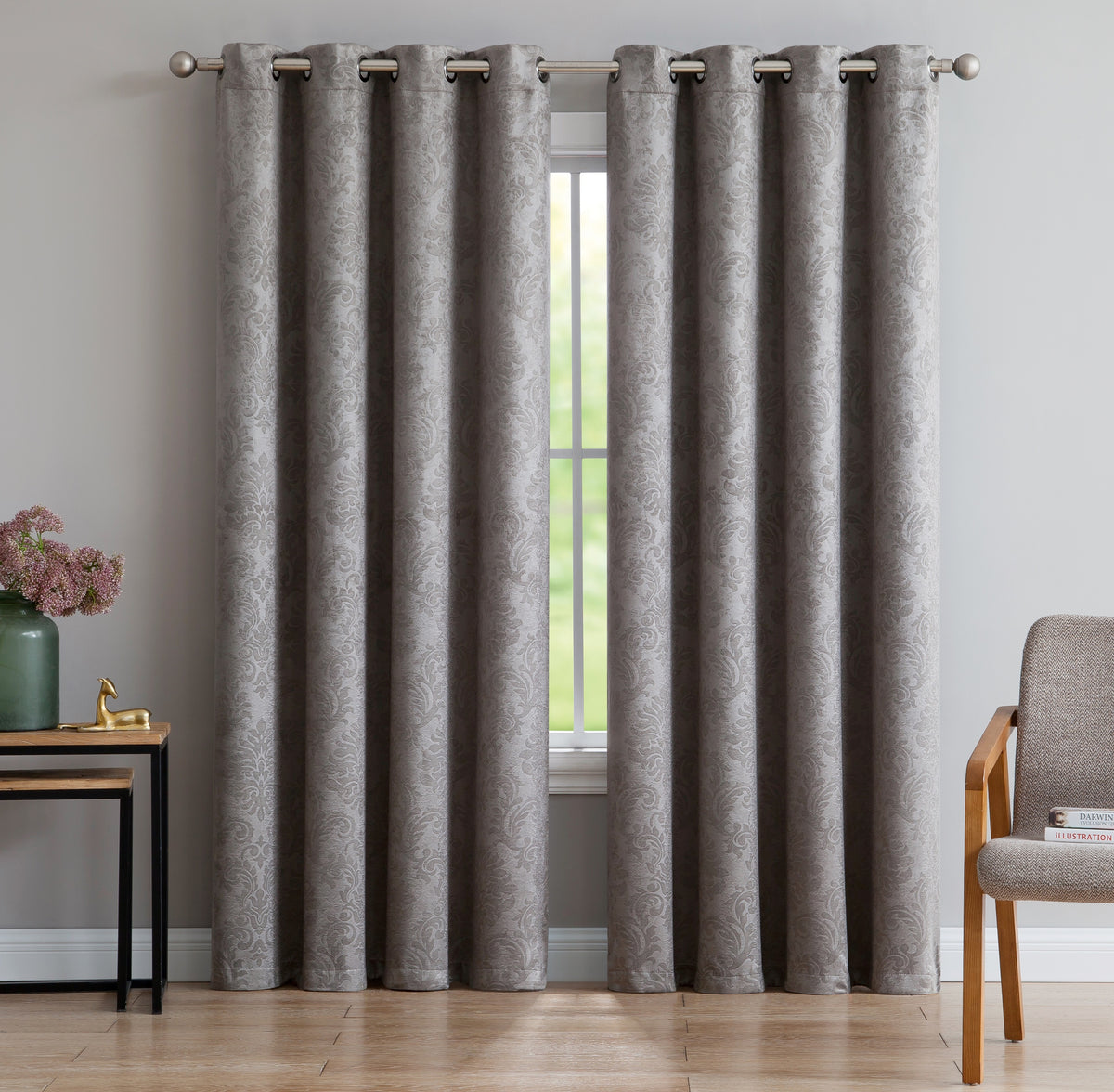 A pair of gray grommet curtains with a subtle damask pattern hanging on a silver rod in a styled room setting with a window, side table, and chair.